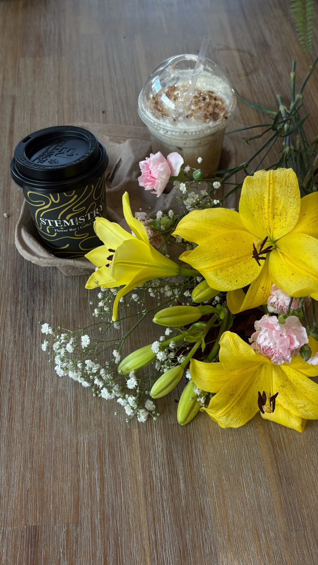 A Stemistry to-go cup and blended iced coffee in a carrier tray beside yellow lilies, pink carnations, and baby's breath